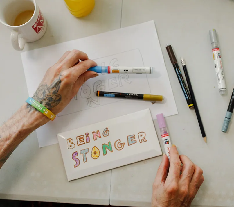 Hands creating artwork with the words 'Being Stronger' during a creative session, representing support for friends and family